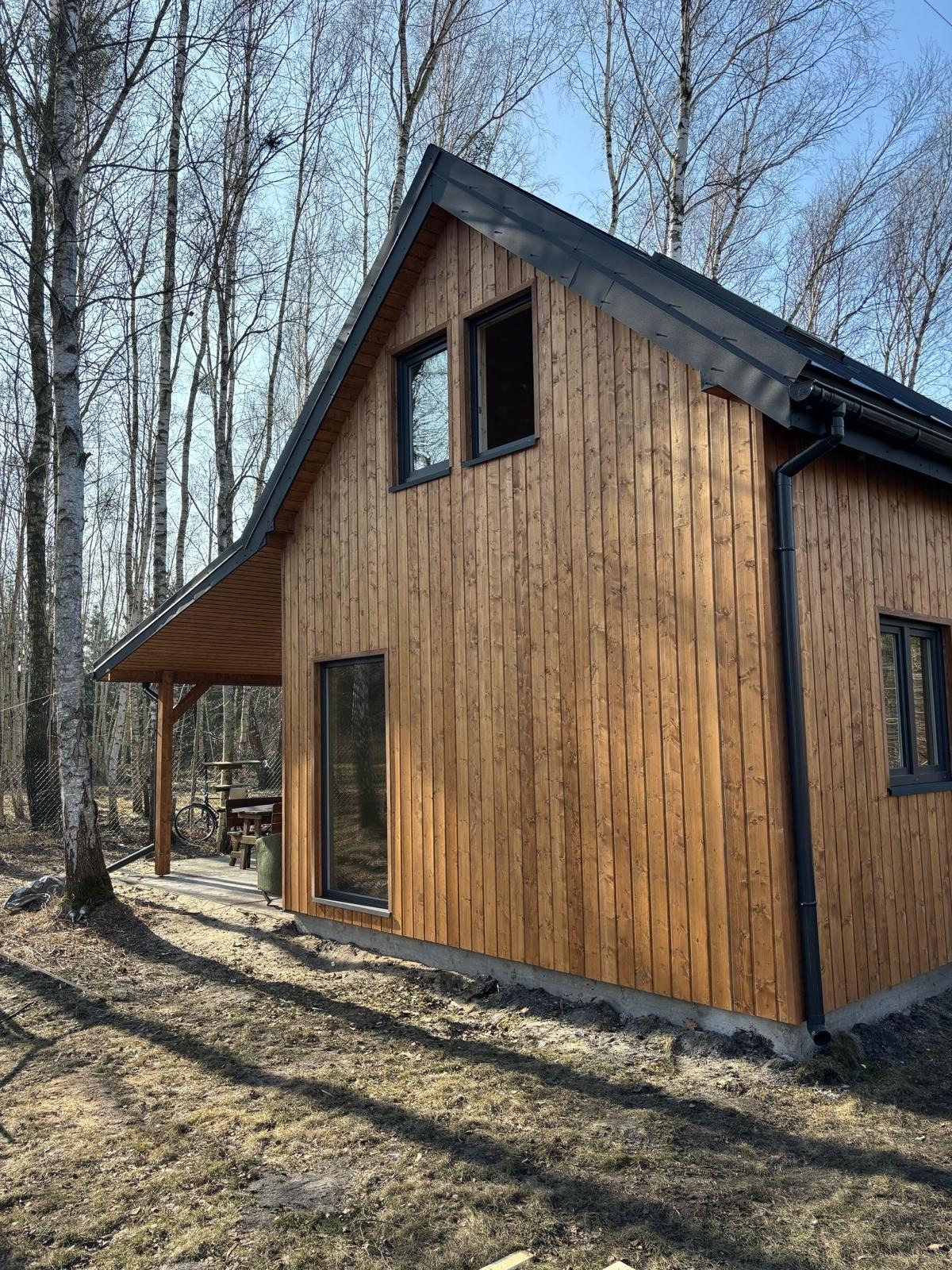 Nowoczesny domek letniskowy z elewacją z jasnego drewna, czarnymi ramami okiennymi i rynnami, otoczony drzewami bez liści w słoneczny dzień.