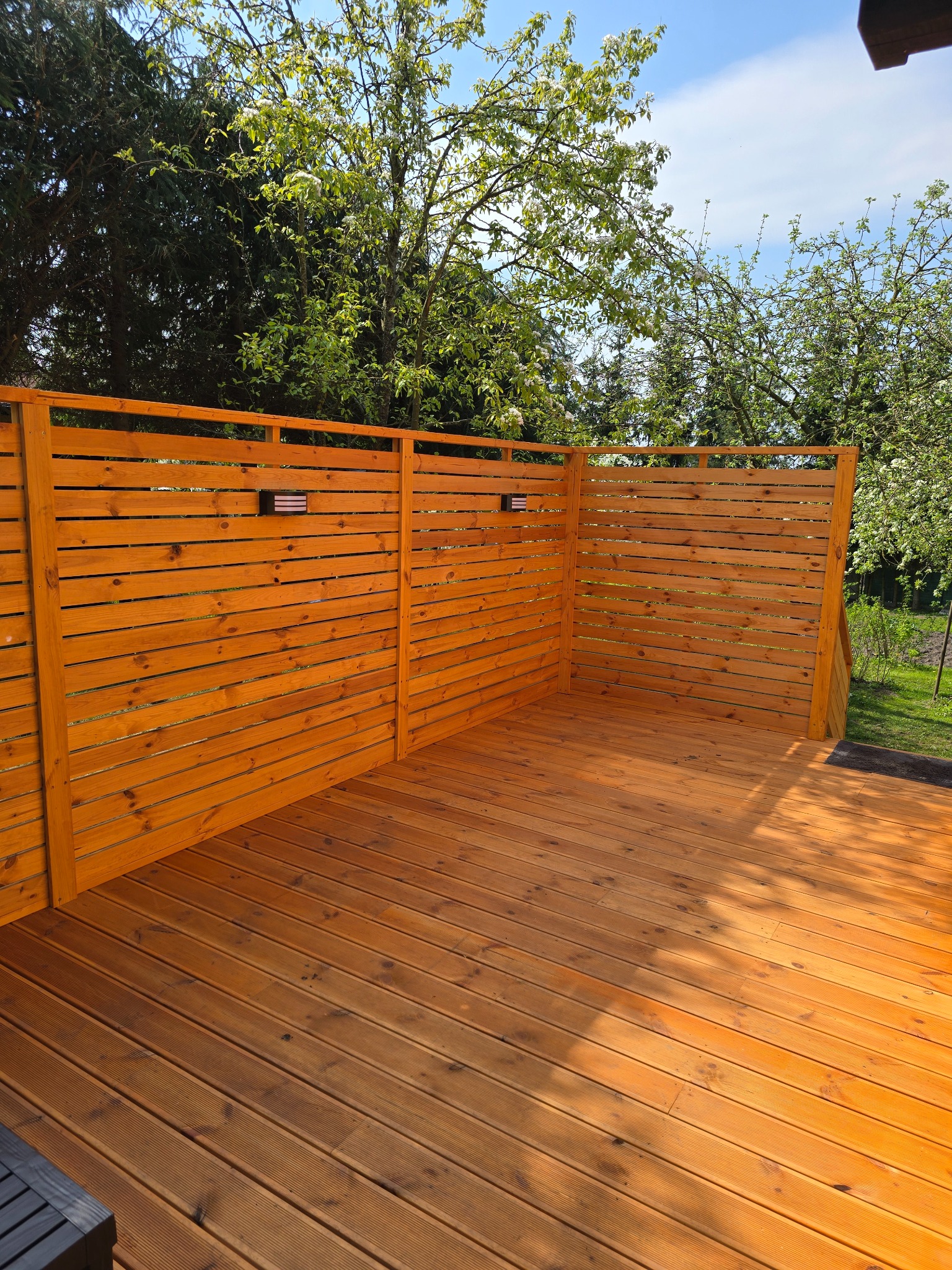 Nowoczesny taras z desek w odcieniach miodu, z drewnianą ażurową ścianką działową z dyskretnym oświetleniem, na tle zieleni drzew i błękitnego nieba.