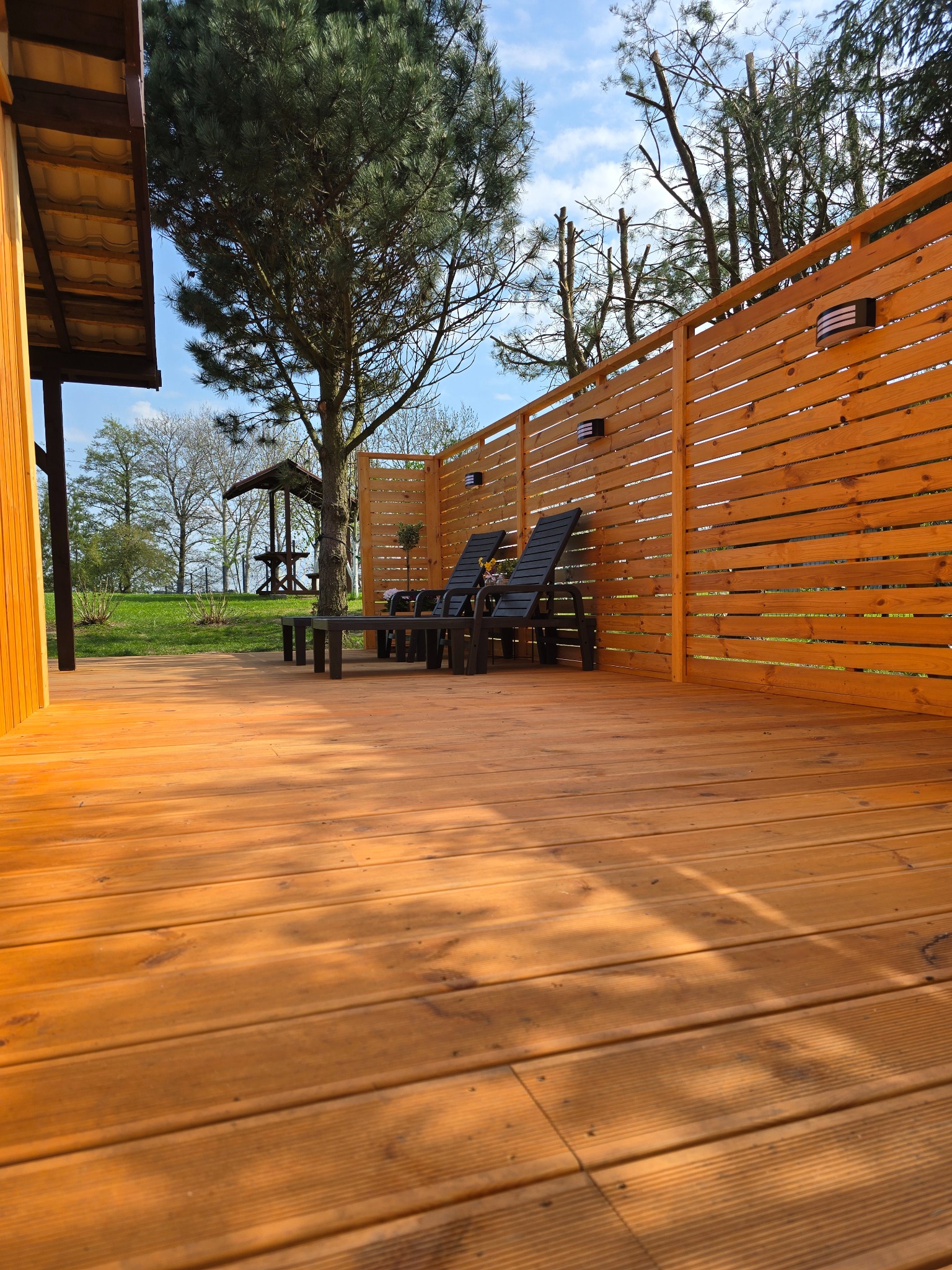 Drewniany taras z leżakami i stolikiem, osłonięty jasnym, poziomym ogrodzeniem z oświetleniem. W tle zielony trawnik i altana, widok w słoneczny dzień.