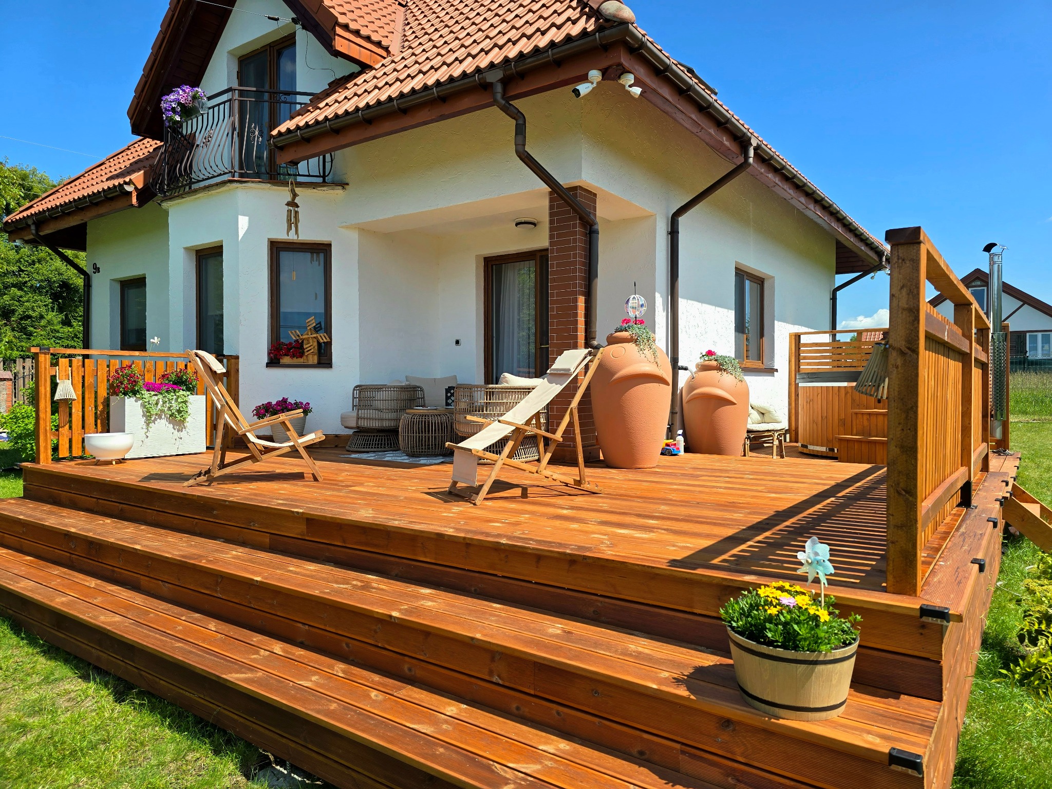 Drewniany taras przy domu z schodami, krzesłami plażowymi, donicami i elementami dekoracyjnymi. Widok na dom z balkonem i zadbanym ogrodem.