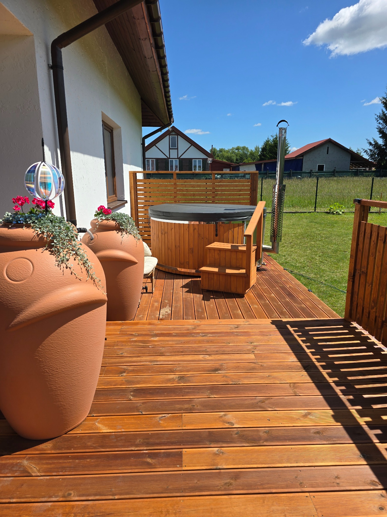 Drewniany taras z jacuzzi i dużymi donicami w kształcie głowy. Elementy drewniane w ciepłym odcieniu brązu. W tle domy i błękitne niebo.