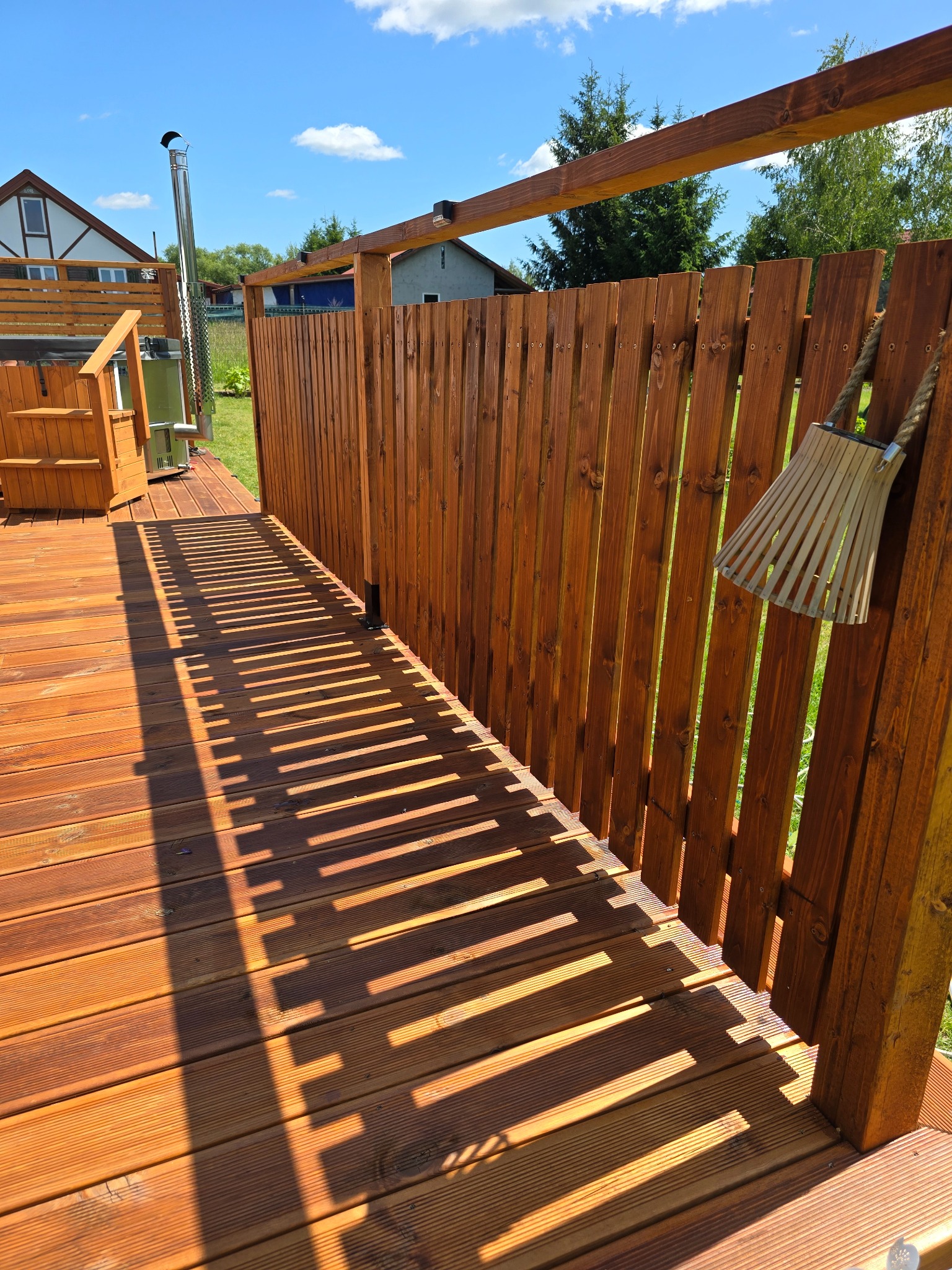 Drewniany taras z jacuzzi, otoczony płotem z desek w ciepłym odcieniu brązu. Na płocie wisi dekoracyjny, ażurowy lampion. Długie cienie podkreślają strukturę desek.