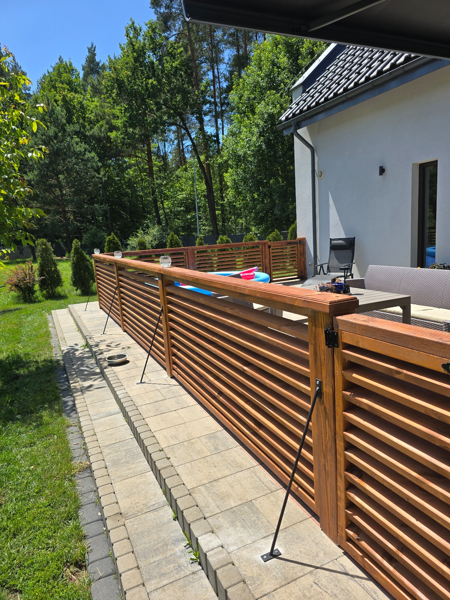 Drewniane, lamelowe ogrodzenie tarasu z ozdobnymi lampionami, wsparte na metalowych podporach. W tle dom z szarą elewacją i zieleń drzew.