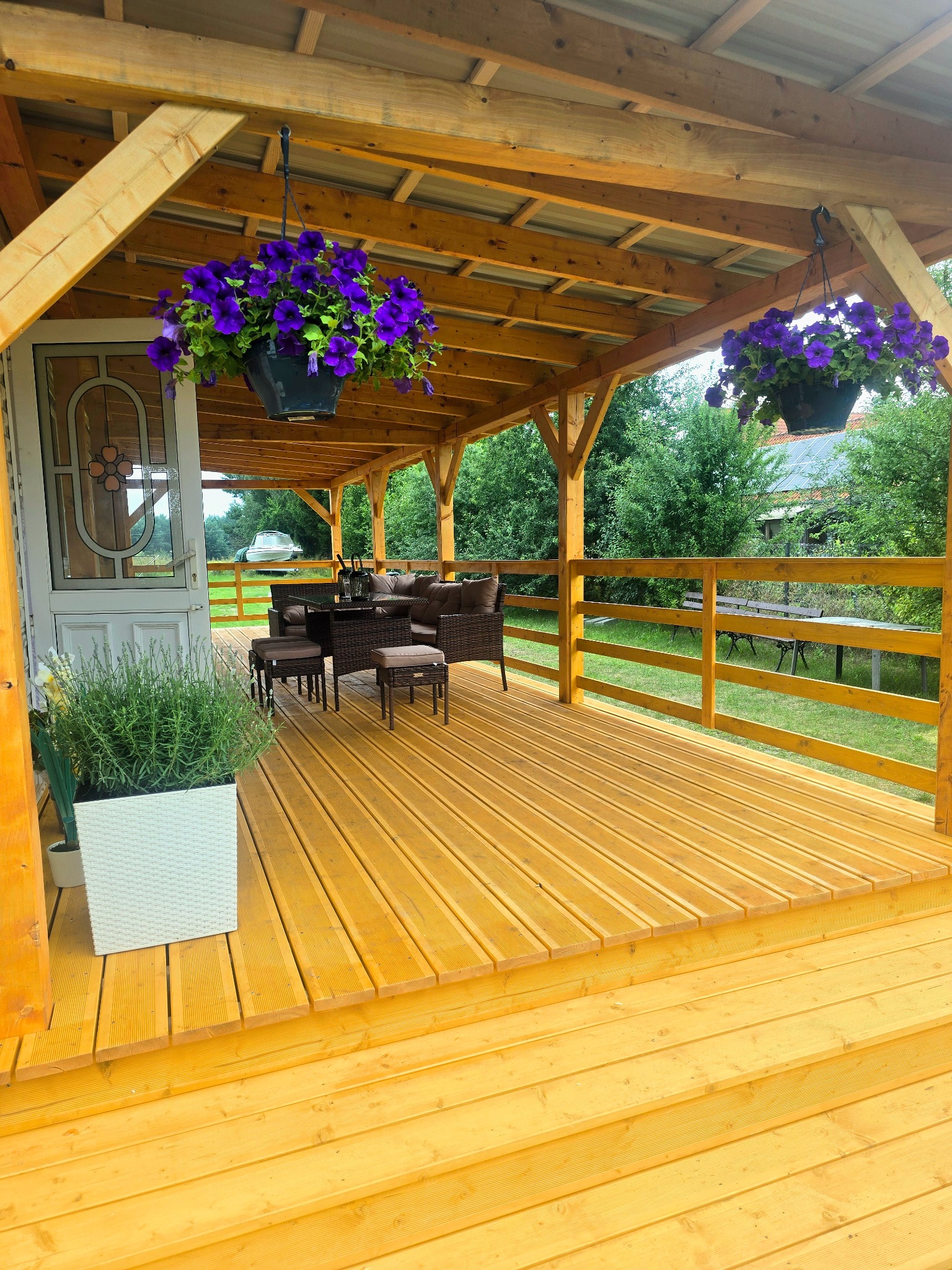 Zadaszony taras drewniany z meblami ogrodowymi i kwiatami w wiszących donicach. Widok na ogród i otoczenie z drewnianą balustradą. Jasne drewno, ciepłe oświetlenie.