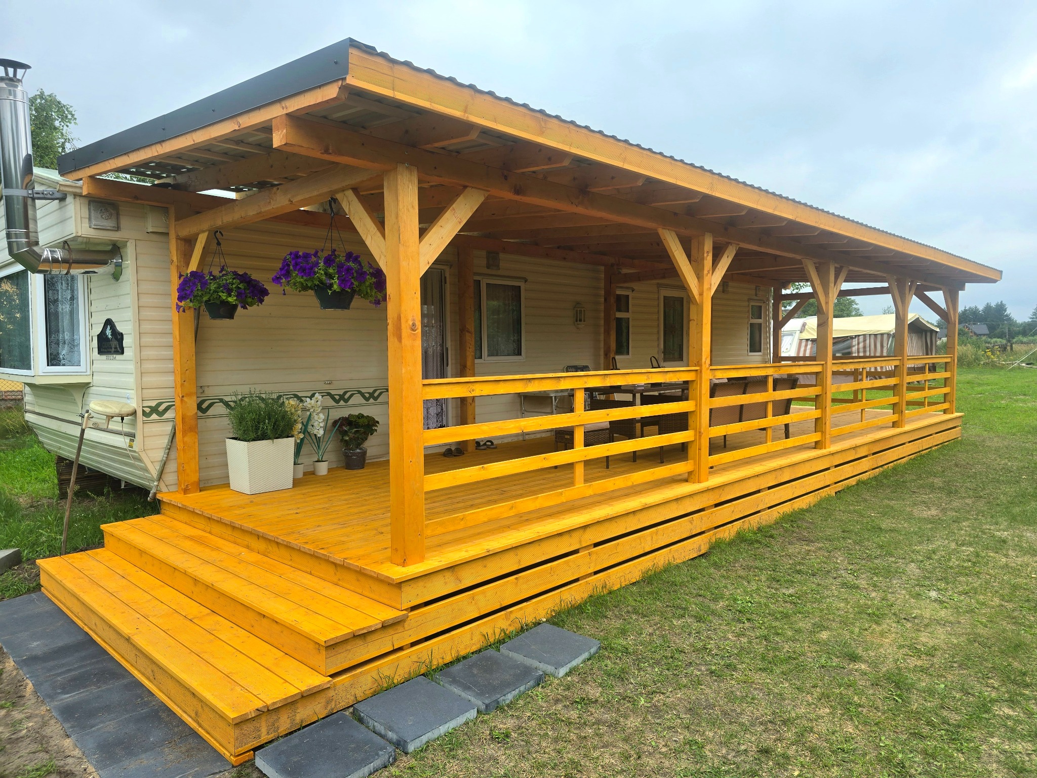 Żółty, drewniany taras z zadaszeniem przyczepiony do mobilnego domu, ozdobiony kwiatami w donicach. Wejście wyłożone kamiennymi płytami.