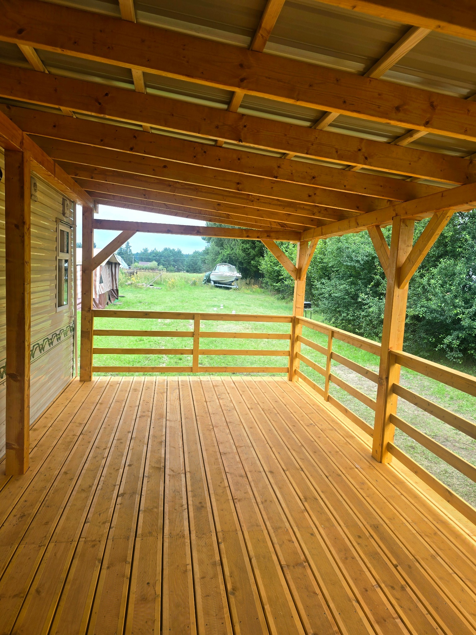Nowy, drewniany taras z balustradą, widok na zielony ogród z łodzią w tle. Jasne drewno, świeżo po budowie. Widok z perspektywy osoby stojącej na tarasie.