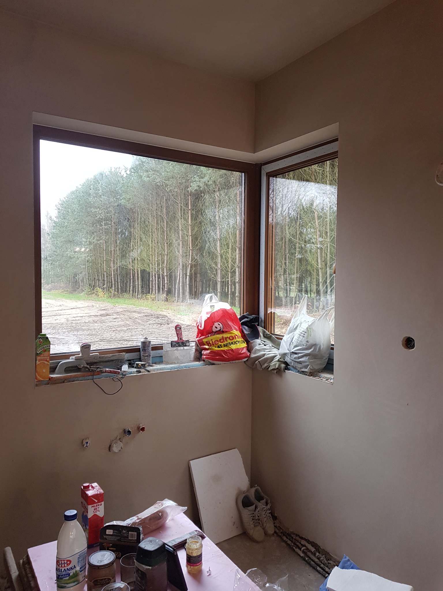 Wnętrze pokoju po nałożeniu tynku gipsowego, widok na okno z narzędziami i artykułami spożywczymi na parapecie, oraz stół z produktami spożywczymi i narzędziami w tle.