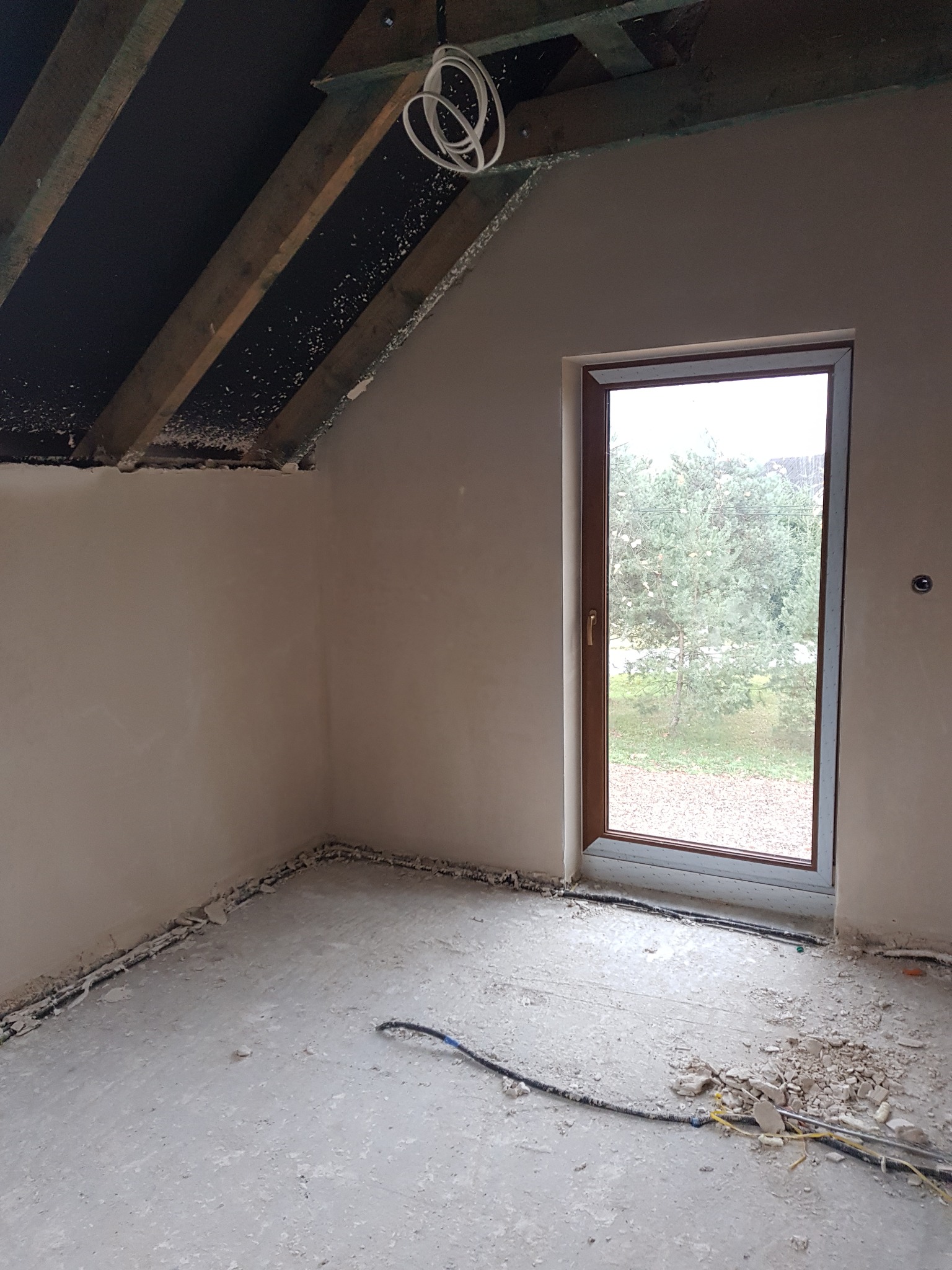 Pokój na poddaszu po nałożeniu wstępnej warstwy tynku, widoczne elementy konstrukcji dachu i okno z widokiem na drzewo.