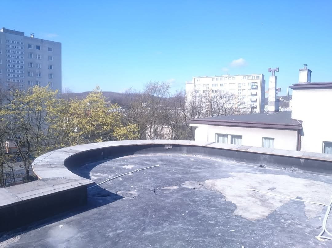 Niedokończony płaski dach z papy na tle budynków mieszkalnych i błękitnego nieba. Widoczne uszkodzenia i wystający kabel. Słoneczny dzień.