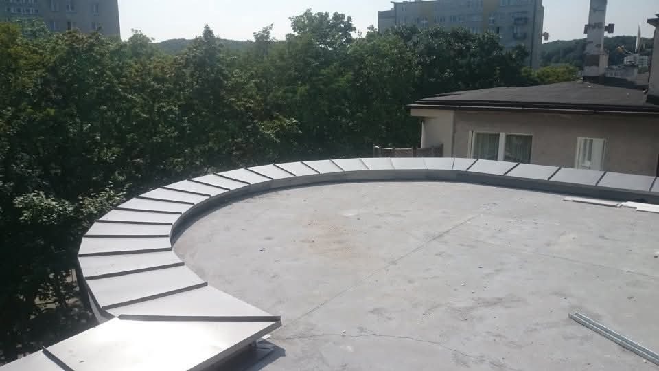 Dach z obróbką blacharską w kształcie łuku, widok z góry. Na dachu widoczne elementy blaszane w odcieniach szarości. W tle zieleń drzew i budynki.