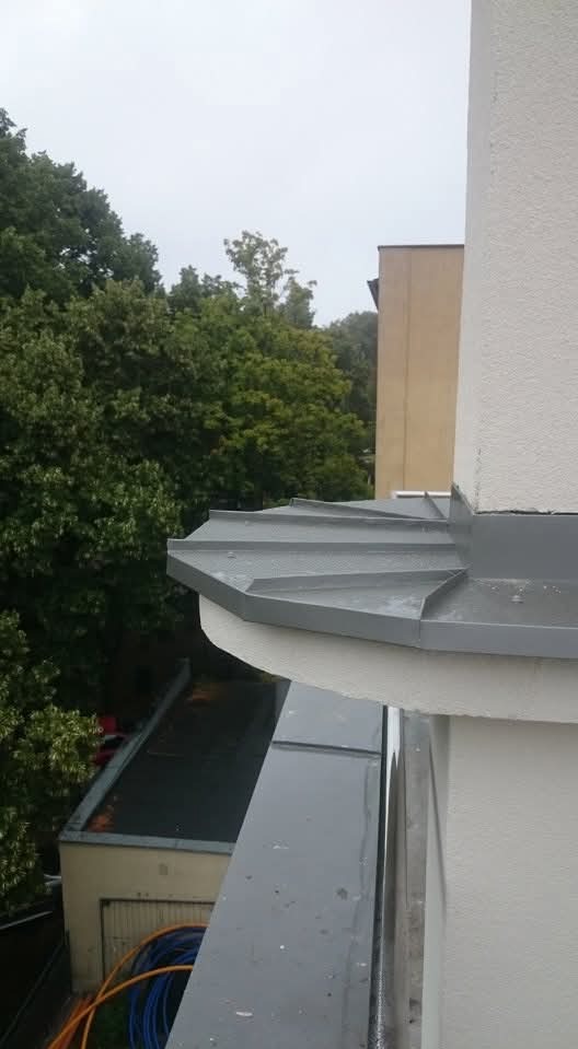 Szare, metalowe wykończenie dachu z widocznymi obróbkami blacharskimi, na tle zieleni drzew i budynku. Widok z góry, z boku balkonu.