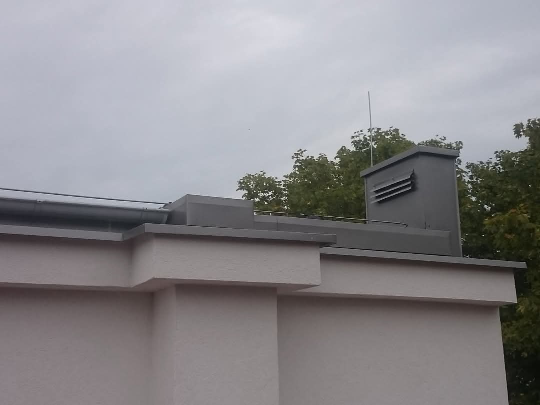 Szary dach z kominem i rynnami na tle pochmurnego nieba i drzew. Widoczne detale obróbek blacharskich i instalacji odgromowej.