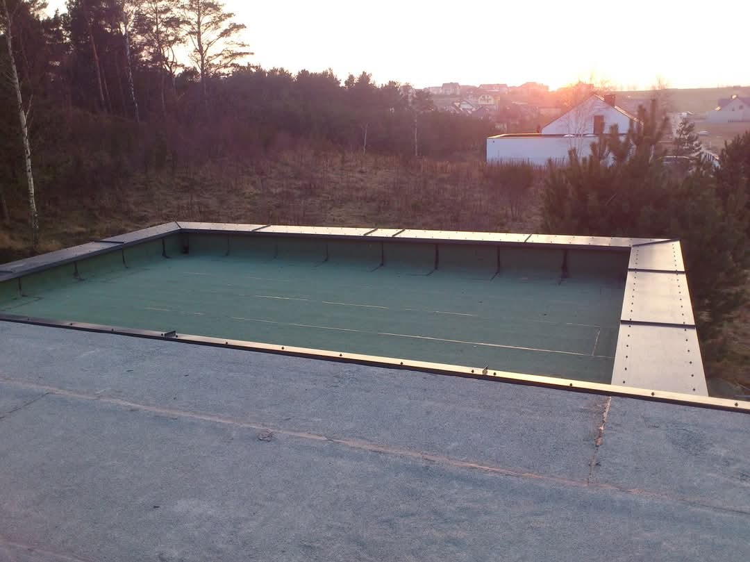 Wykończony dach płaski z obróbkami blacharskimi w Gdyni, widok z góry na tle zieleni i zabudowań w oddali, ciepłe światło zachodzącego słońca.