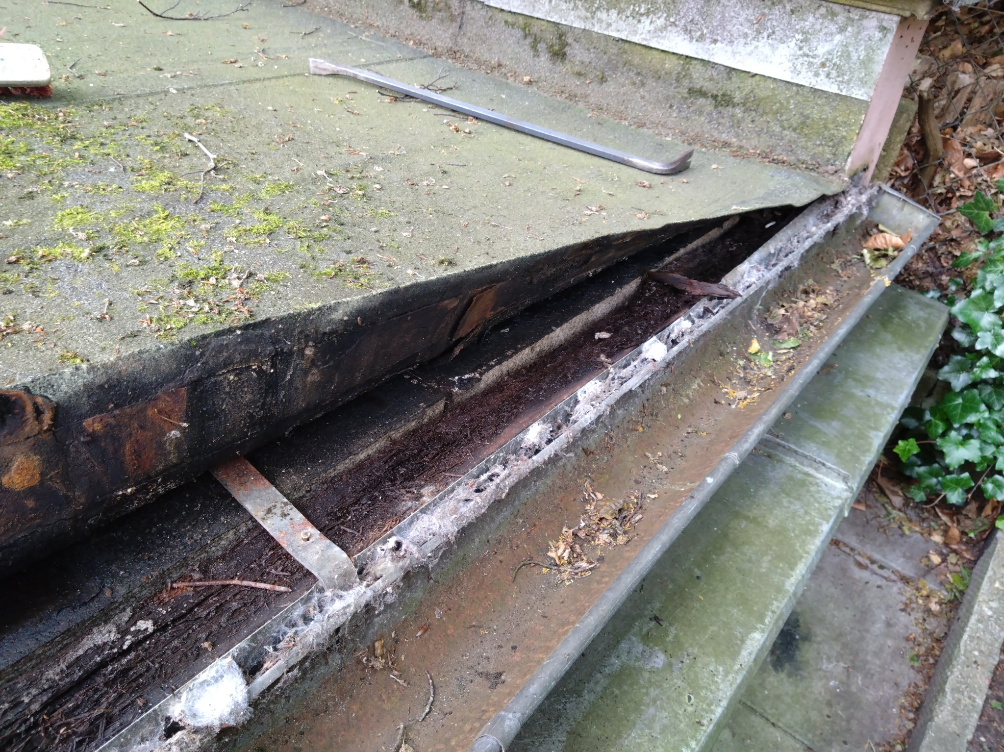 Otwarta, zardzewiała rynna z resztkami liści i mchu, widoczna spod uszkodzonego dachu. Na dachu leży metalowy łom.