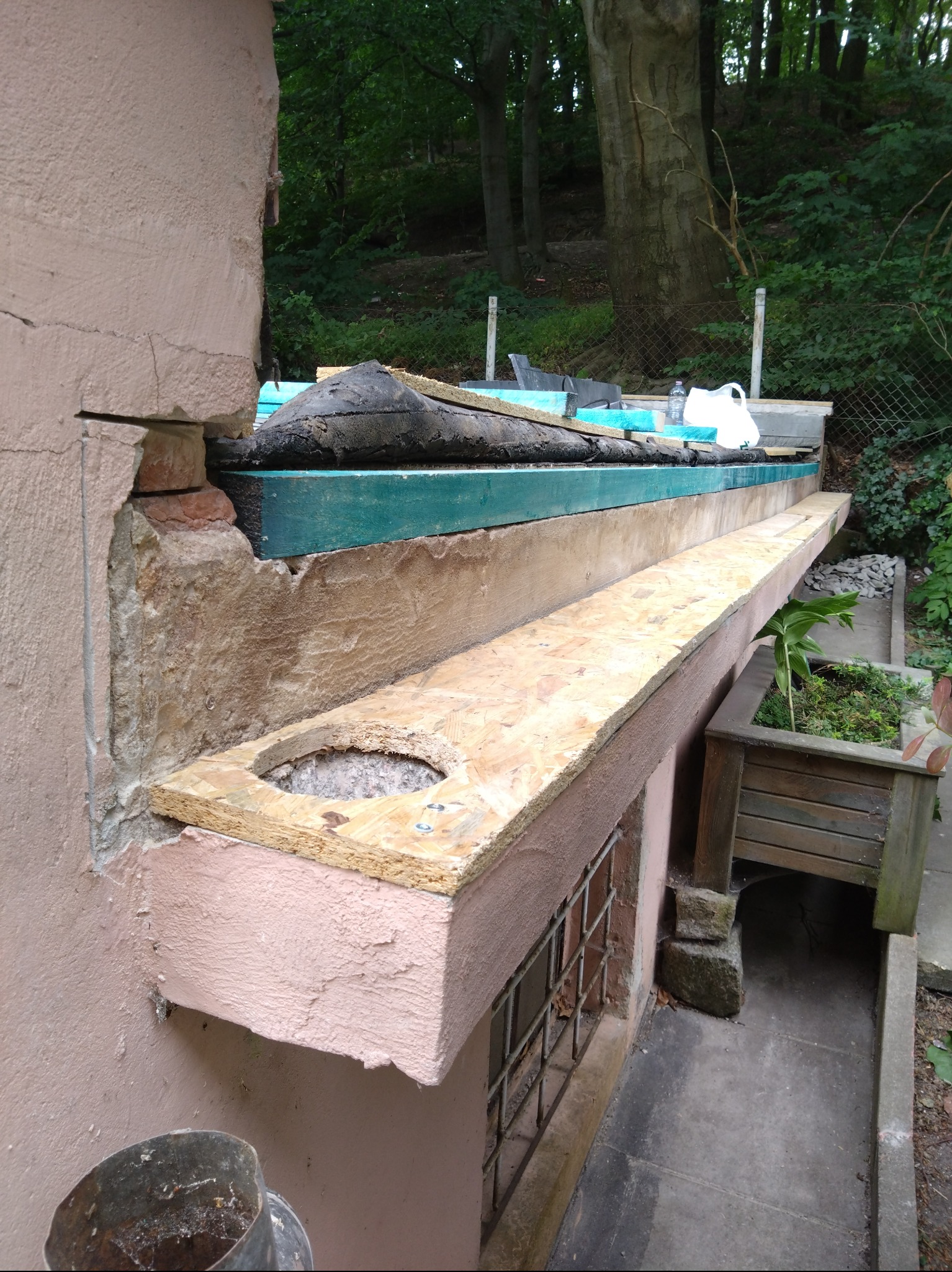 Fragment dachu w trakcie naprawy/uszczelniania z widoczną konstrukcją i materiałami izolacyjnymi, na tle zieleni.