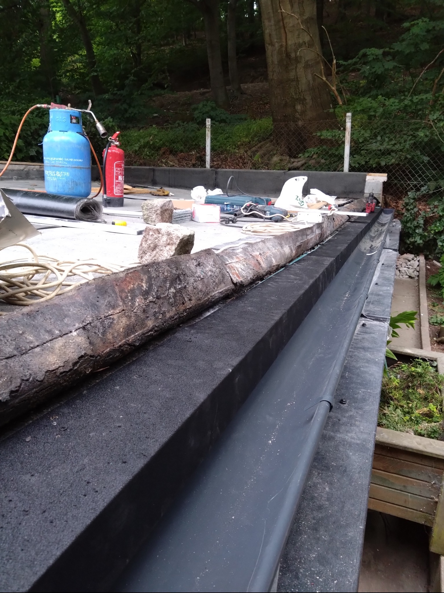 Prace dekarskie na płaskim dachu: widoczna nowa, grafitowa rynna, narzędzia, butla gazowa i pozostałości starego pokrycia. Prace w toku, otoczenie leśne.