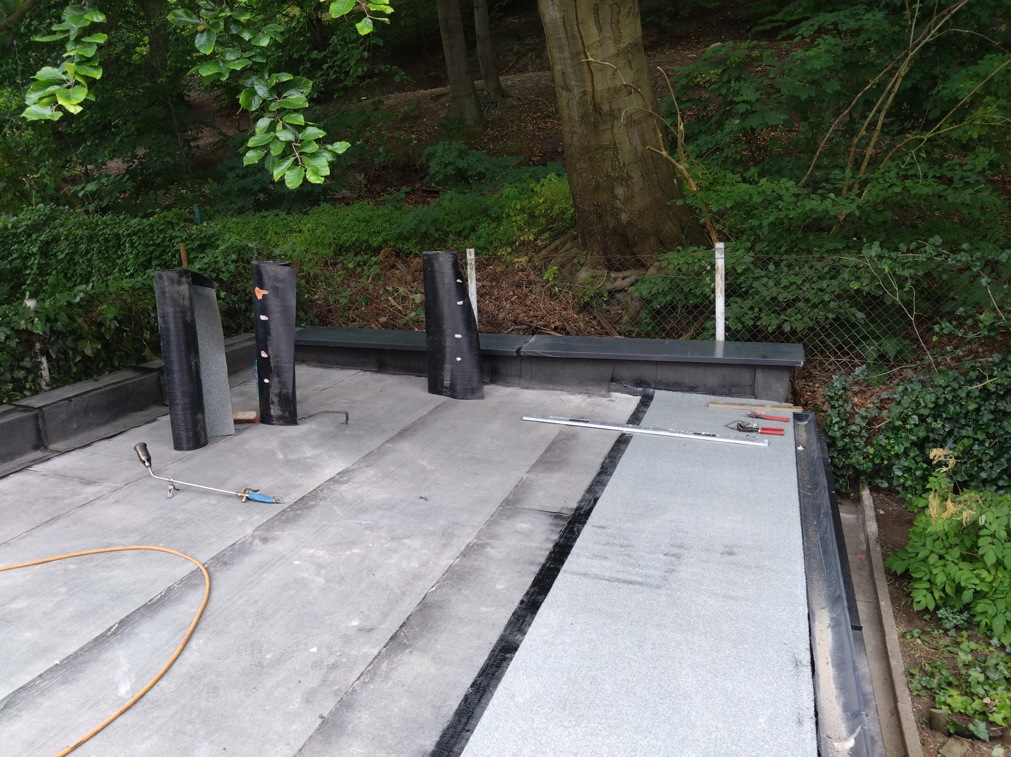 Prace na dachu płaskim: papa termozgrzewalna zgrzewana palnikiem, widoczne narzędzia i pionowe elementy, tło zieleni.