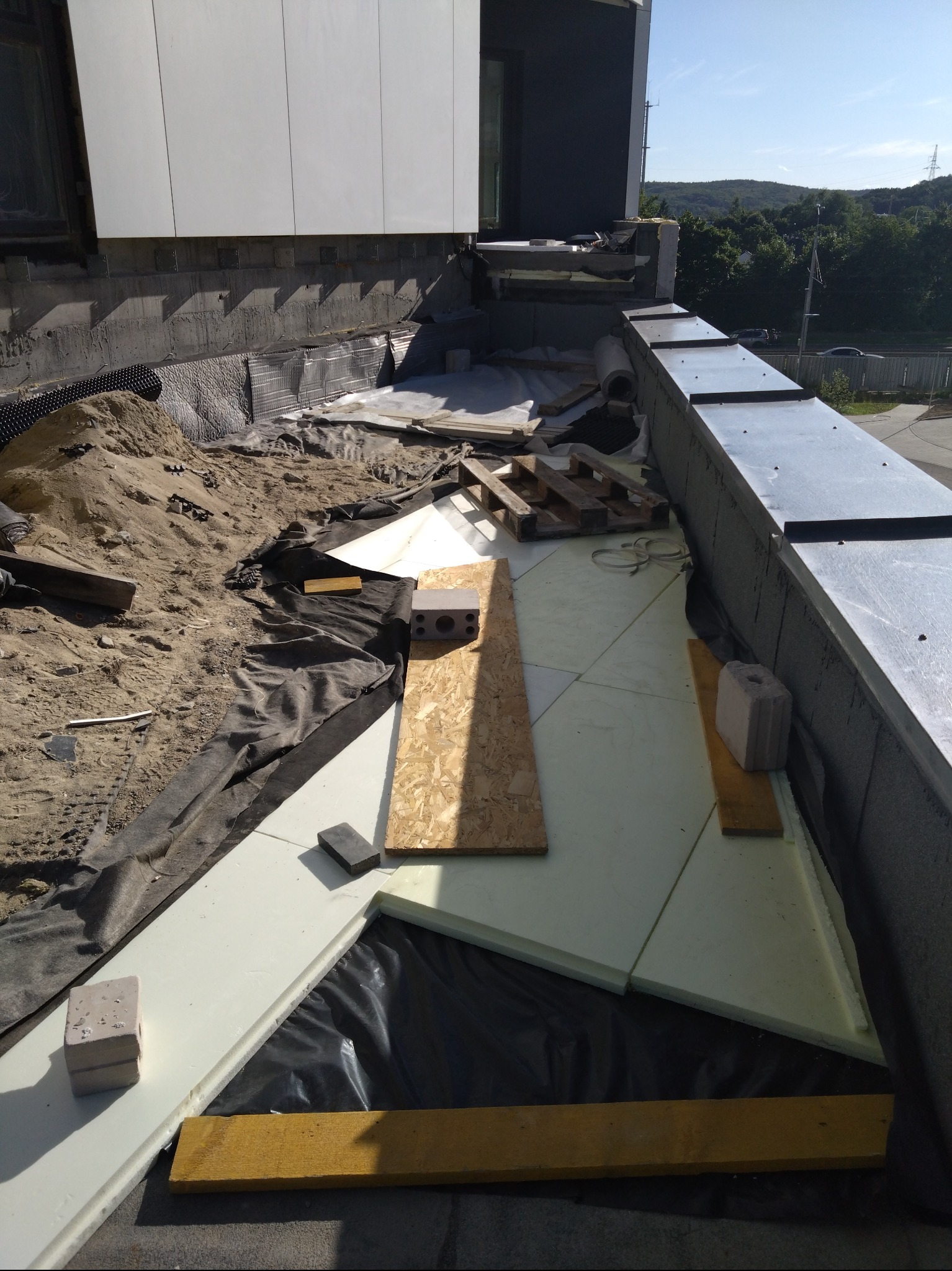 Montaż izolacji termicznej dachu płaskiego na nowoczesnym budynku. Widoczne płyty styropianowe, membrana, piasek i obróbki blacharskie w słoneczny dzień.