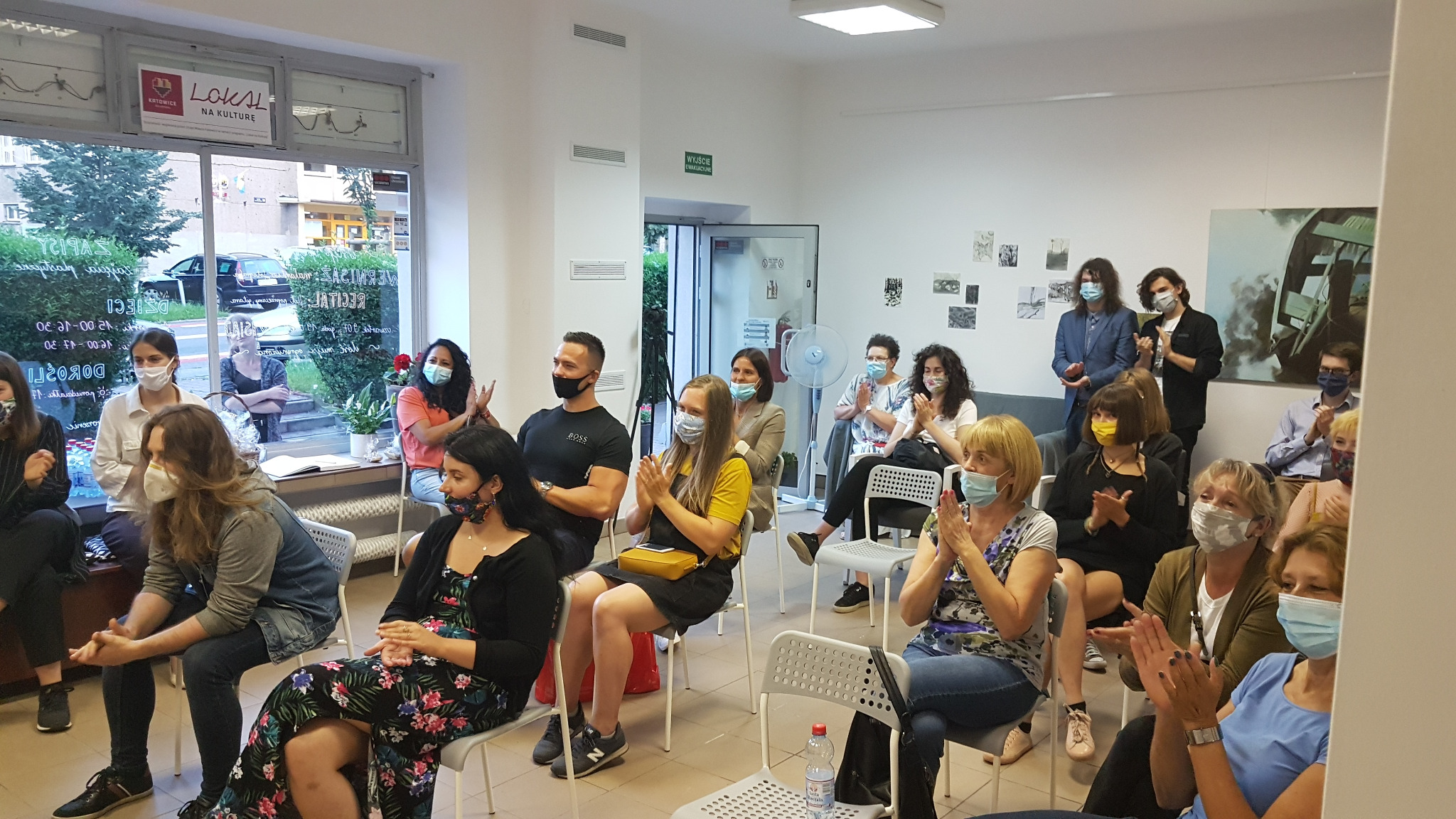 Wernisaż w Galerii Alabastro: publiczność w maseczkach siedzi na krzesłach i klaszcze po recitalu. W tle widoczne obrazy na ścianie i okno z napisem 'Lokal na Kulturę'.