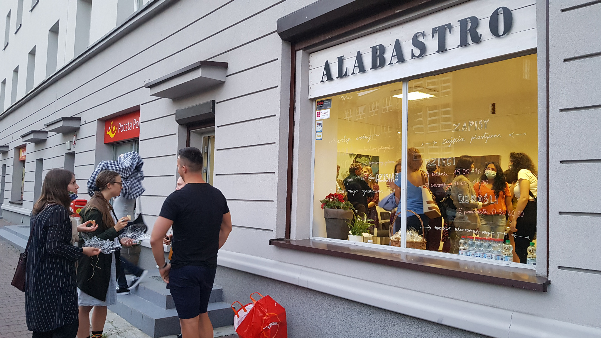 Fasada budynku z szyldem 'ALABASTRO', przez okno widoczni ludzie wewnątrz, na chodniku grupa osób z przekąskami i napojami, obok wejścia torba zakupowa.
