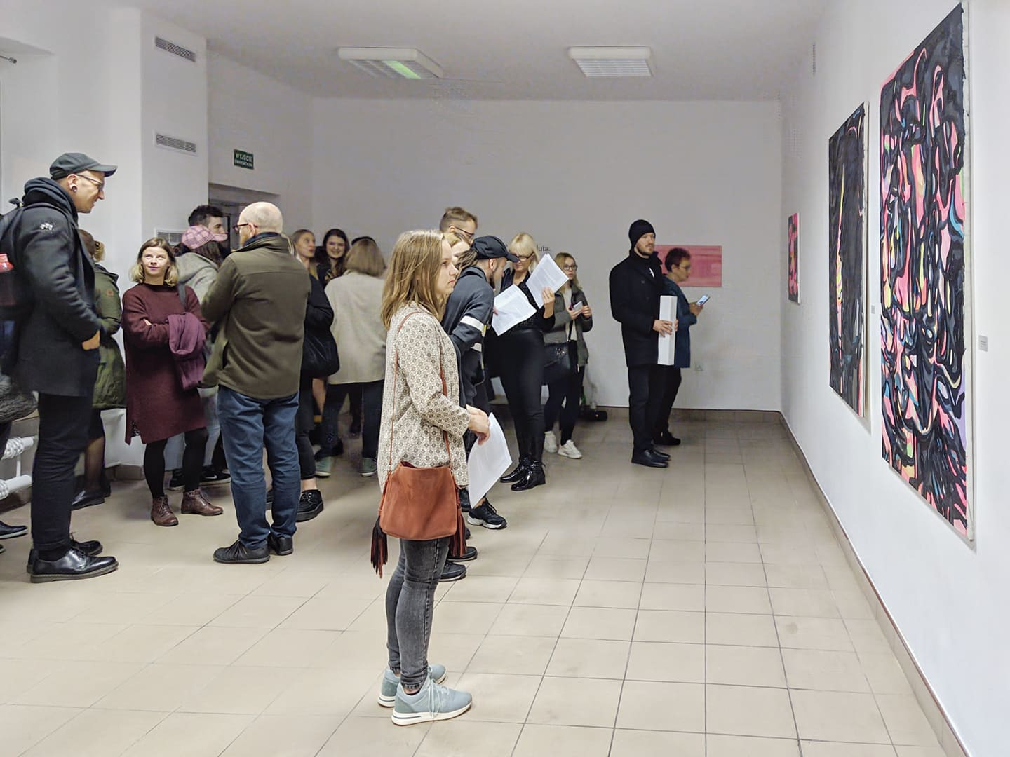 Wernisaż w galerii: grupa ludzi ogląda abstrakcyjne obrazy w czarnych ramach, wiszące na białej ścianie, widoczne białe płytki podłogowe i oświetlenie sufitowe.