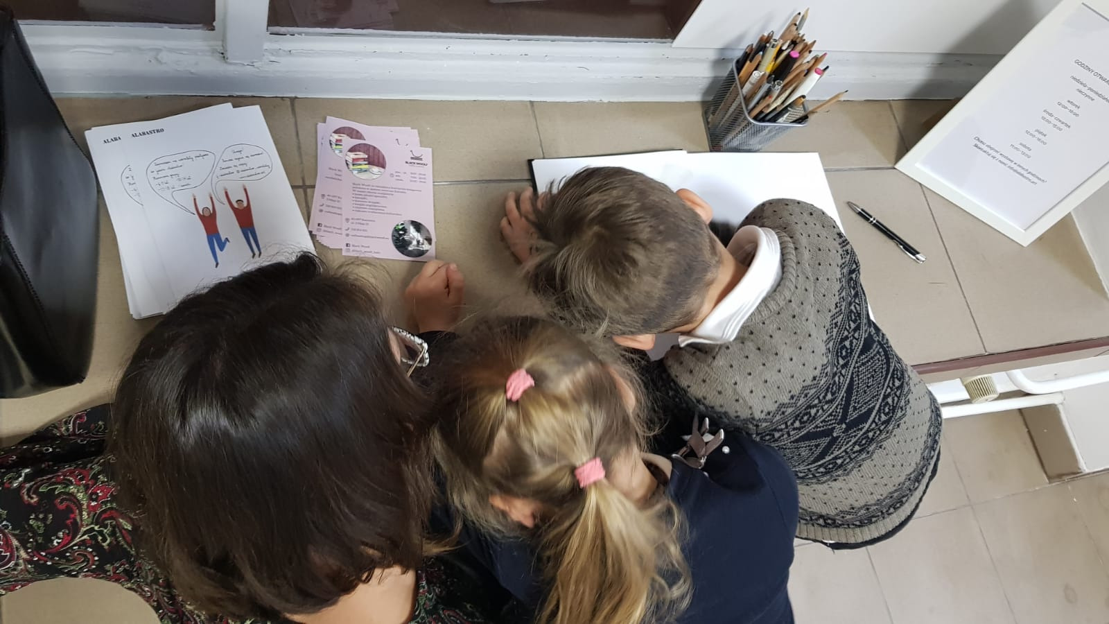 Wernisaż w Galerii Alabastro: kobieta i dwoje dzieci pochyleni nad rysunkami i kartkami informacyjnymi, widok z góry na stolik z ołówkami i godzinami otwarcia galerii.