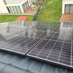 Panele słoneczne na dachu domu jednorodzinnego, widok z góry na czarne moduły z odbiciami nieba i otoczenia, w tle ogród z elementami architektury.