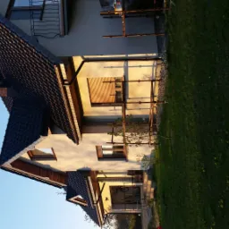 Dom jednorodzinny w trakcie prac elewacyjnych, widoczne rusztowania przy ścianie, świeżo położony tynk oraz fragment trawnika z żółtymi kwiatami.