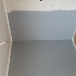 Stanisław Napierała - Świeżo pomalowana na szaro ściana w pomieszczeniu, widoczne wystające kable elektryczne i zabrudzona podłoga z resztkami farby, wałek do malowania w rogu.