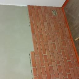 Stanisław Napierała - Fragment ściany z częściowo nałożoną okładziną z czerwonej cegły, widoczna puszka elektryczna, obok ściana pokryta szarą gładzią, na podłodze wiadro z zaprawą.