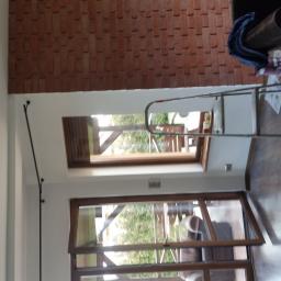 Stanisław Napierała - Wnętrze z drewnianą podłogą i ceglaną ścianą, widoczne okno dachowe i okno balkonowe, oraz drabina oparta o ścianę, sugerująca prace wykończeniowe lub naprawcze.