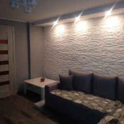 Nowoczesny salon z dekoracyjną ścianą z białej cegły, podświetloną punktowymi lampami sufitowymi, szarą sofą z narzutą w liście oraz drewnianą podłogą.