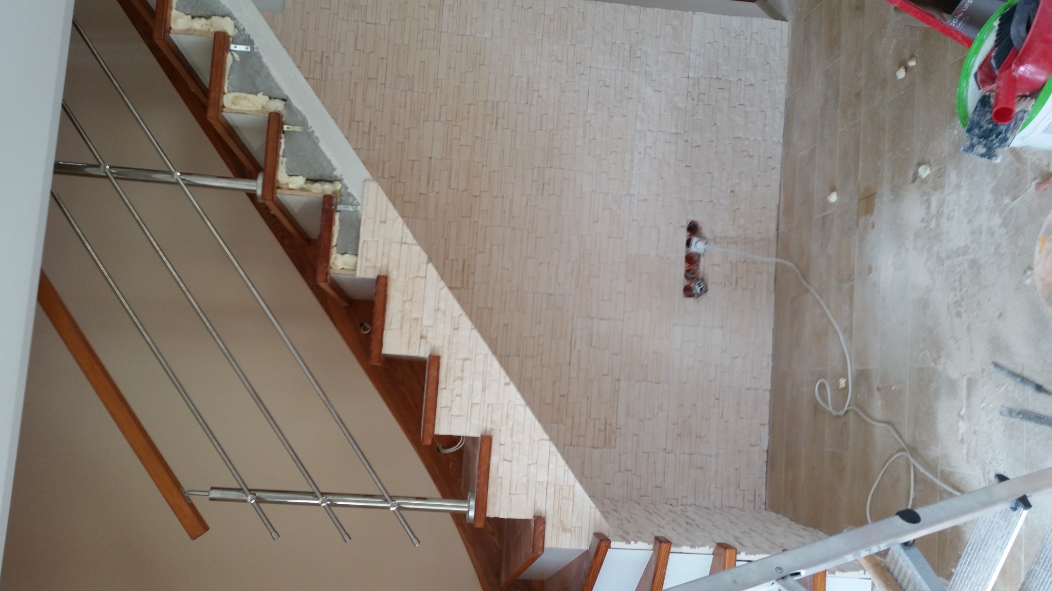 Montaż drewnianych schodów z metalową balustradą, widoczne ocieplenie pianką montażową, ściana wyłożona dekoracyjną cegłą, podłoga w trakcie prac wykończeniowych.