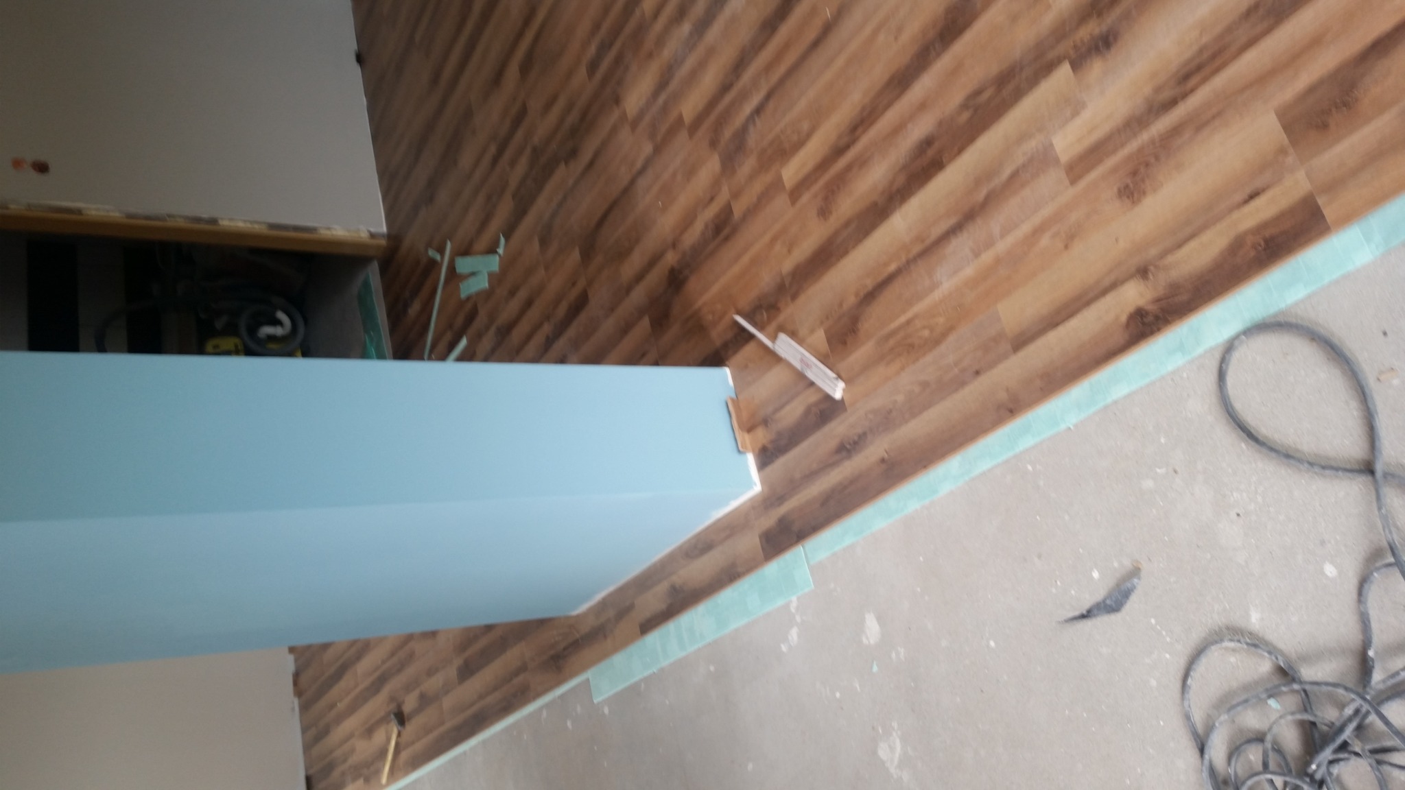 Układanie paneli podłogowych z imitacją drewna, widoczna niebieska ściana działowa, fragment kabla elektrycznego i listwy przypodłogowe w trakcie montażu.