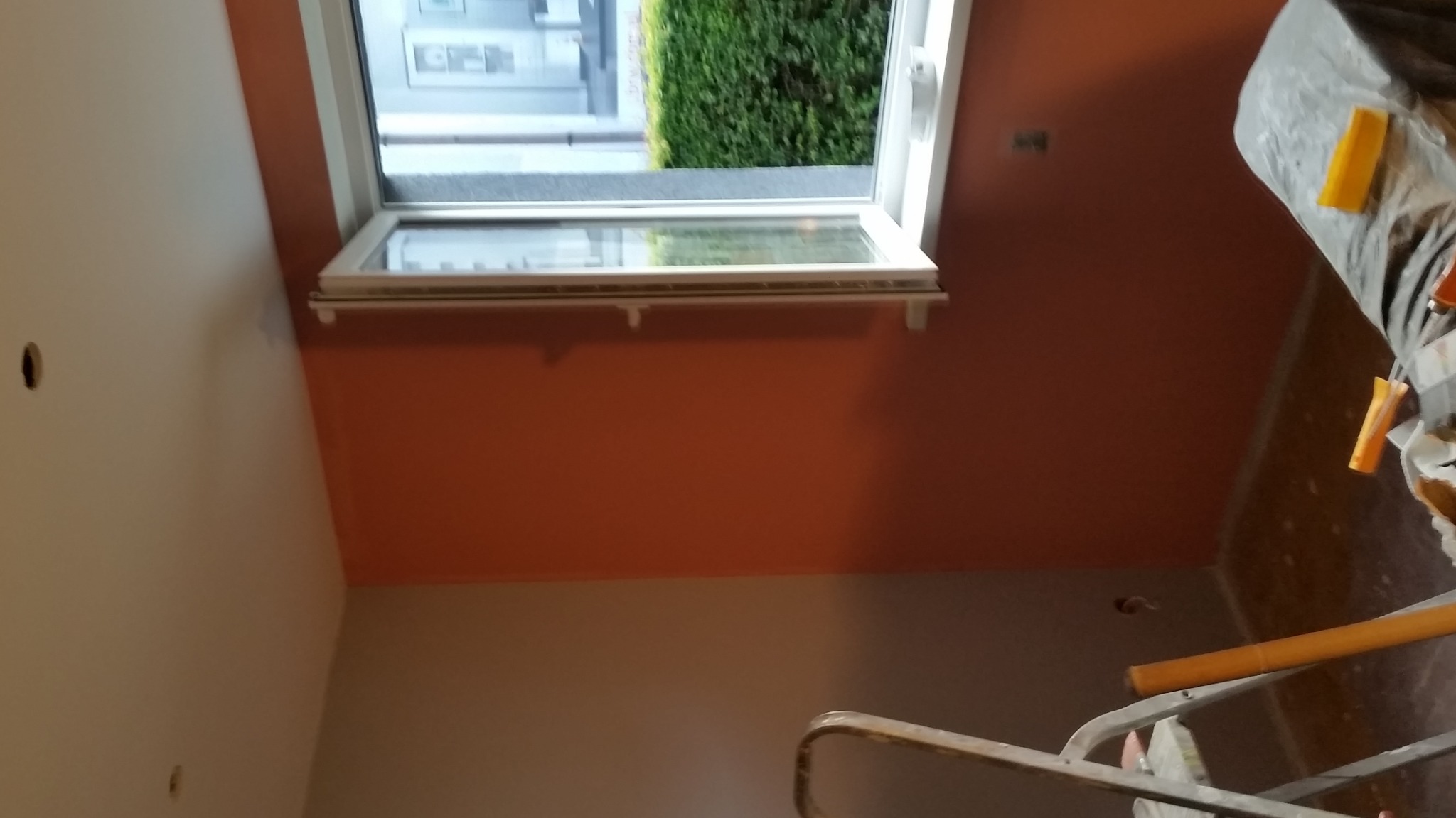 Ściana pomalowana na pomarańczowo w Żninie, widoczne okno dachowe i fragment drabiny, prace wykończeniowe w trakcie.