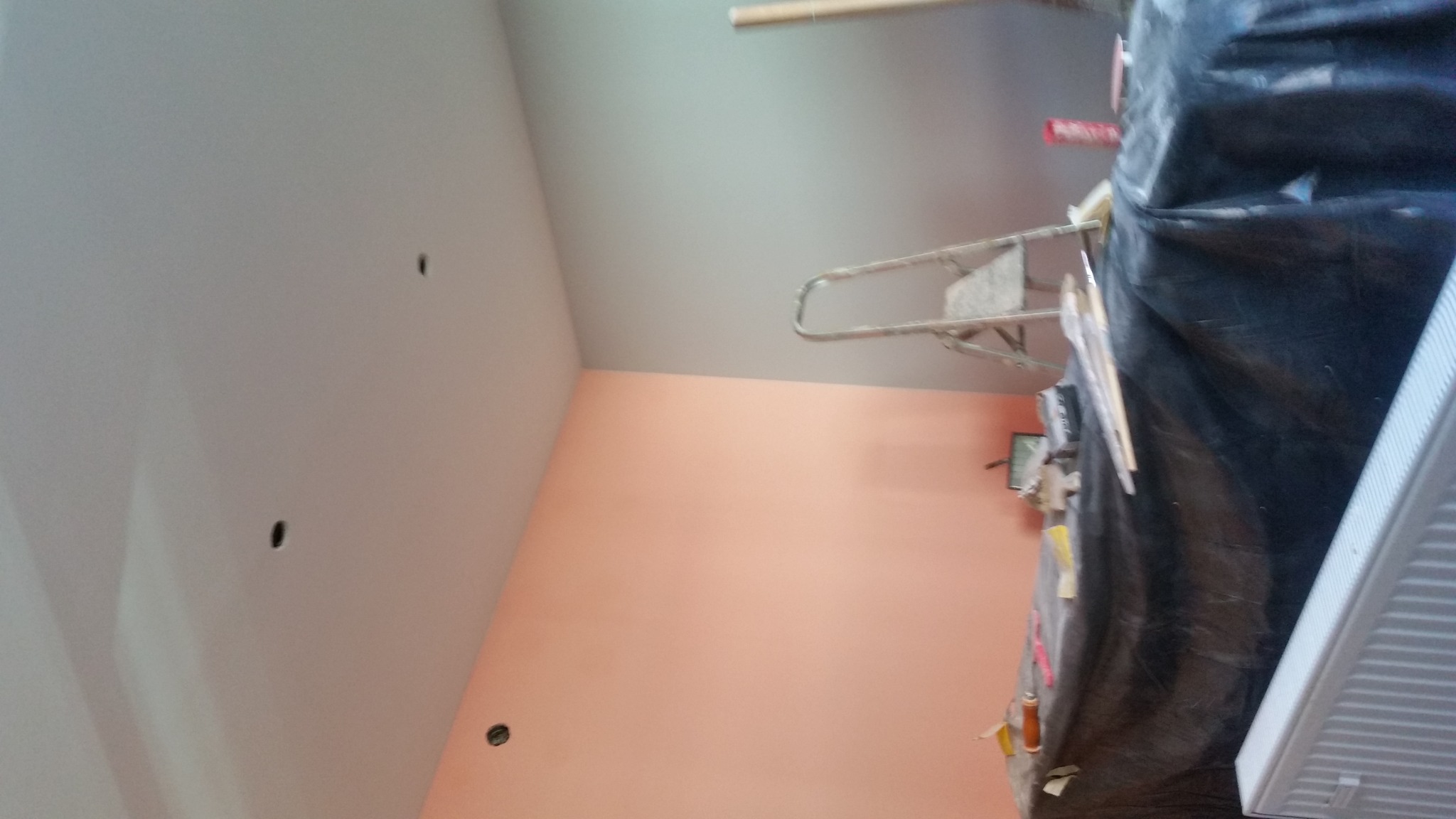 Wnętrze pokoju w trakcie prac przygotowawczych, z widoczną drabiną, zakrytymi meblami folią ochronną, pomalowaną na łososiowy kolor ścianą i surowym sufitem z widocznymi otworami.