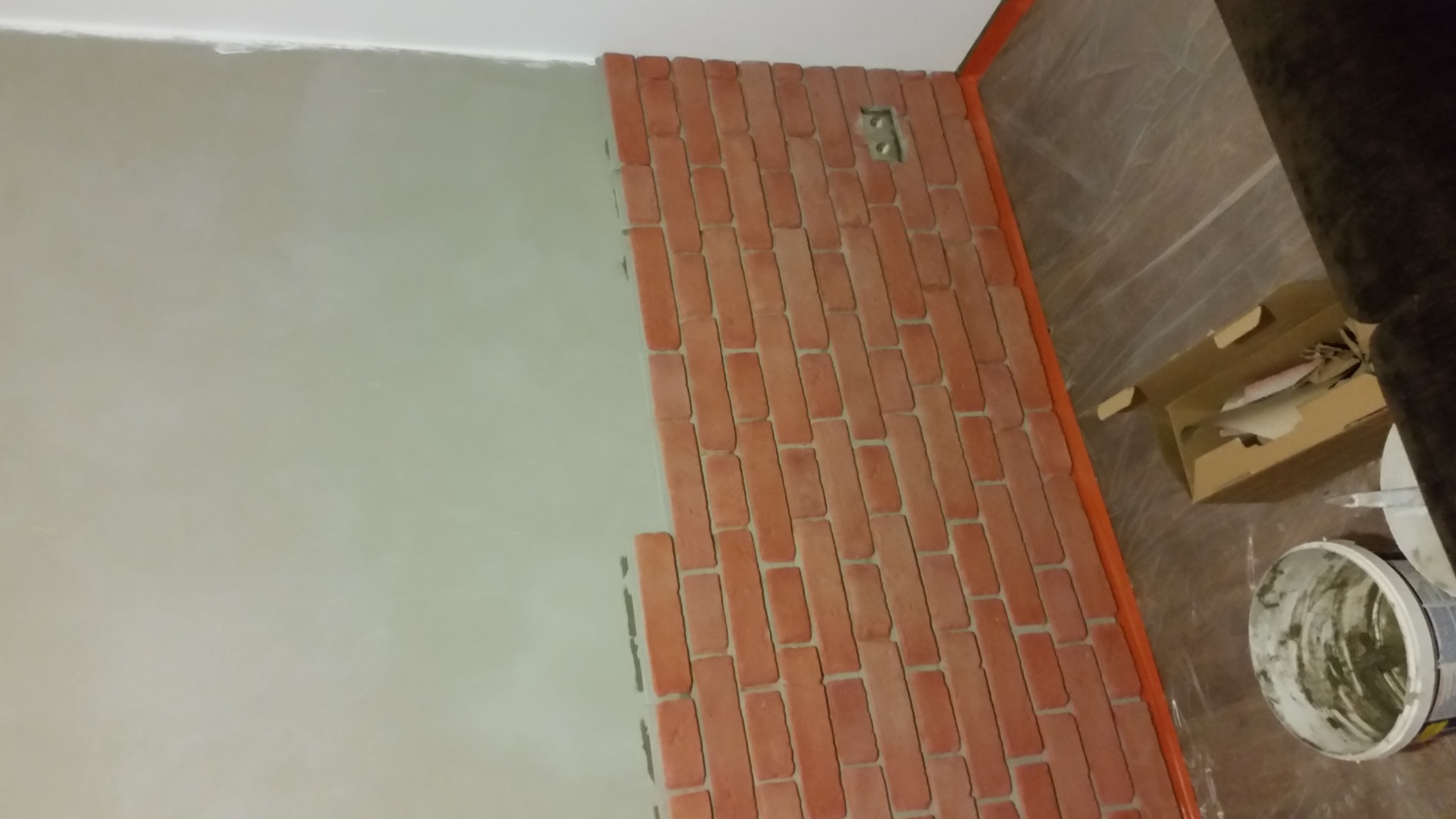 Fragment ściany z częściowo nałożoną okładziną z czerwonej cegły, widoczna puszka elektryczna, obok ściana pokryta szarą gładzią, na podłodze wiadro z zaprawą.