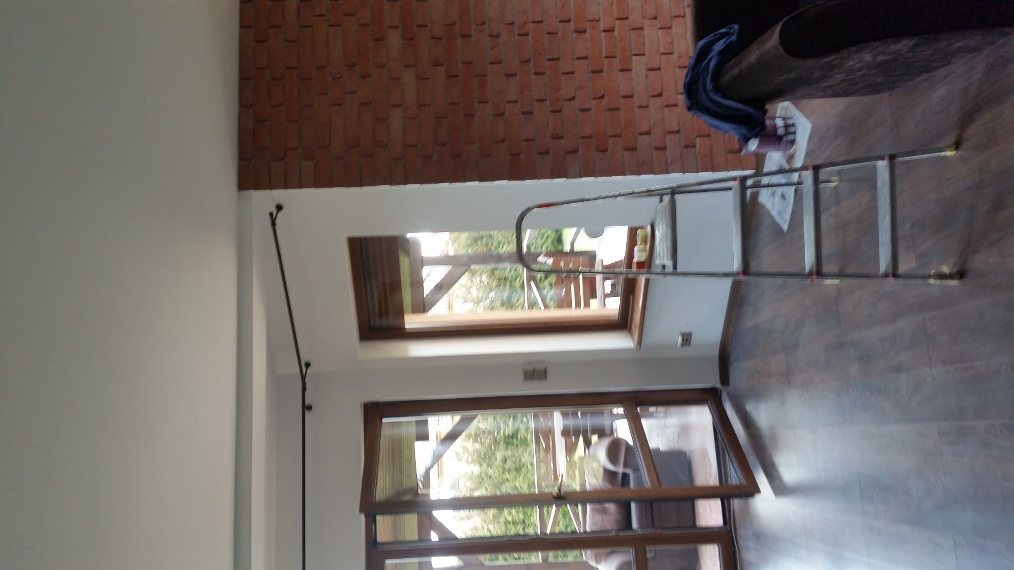 Wnętrze z drewnianą podłogą i ceglaną ścianą, widoczne okno dachowe i okno balkonowe, oraz drabina oparta o ścianę, sugerująca prace wykończeniowe lub naprawcze.