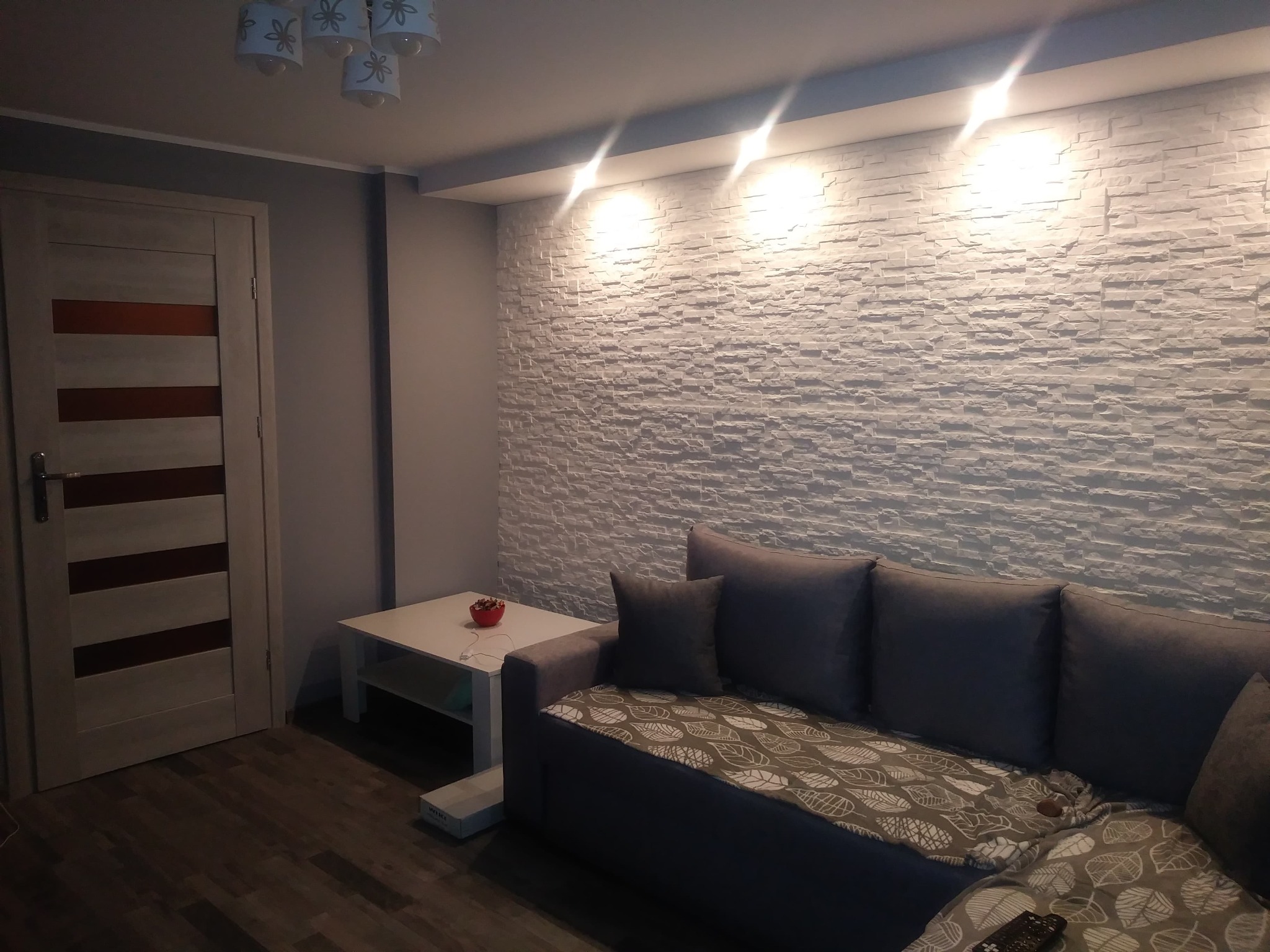 Nowoczesny salon z dekoracyjną ścianą z białej cegły, podświetloną punktowymi lampami sufitowymi, szarą sofą z narzutą w liście oraz drewnianą podłogą.