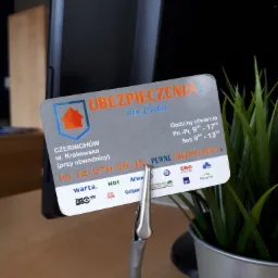 Wizytówka firmy ubezpieczeniowej Czernichów, z adresem, numerem telefonu i logotypami różnych towarzystw, umieszczona w metalowym uchwycie na biurku obok rośliny doniczkowej.