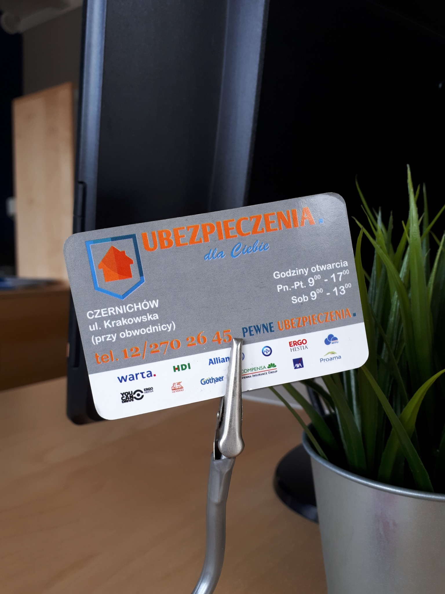 Wizytówka firmy ubezpieczeniowej Czernichów, z adresem, numerem telefonu i logotypami różnych towarzystw, umieszczona w metalowym uchwycie na biurku obok rośliny doniczkowej.