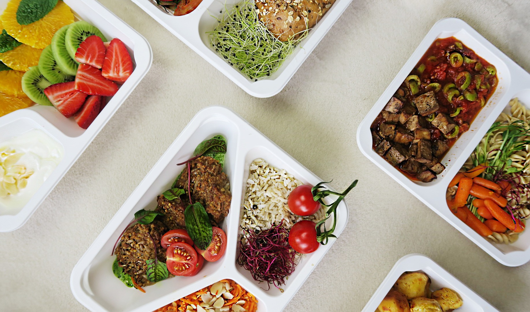 Różnorodne posiłki w białych, plastikowych pojemnikach z podziałem na sekcje, zawierające m.in. owoce, warzywa, mięso i kasze, ułożone na jasnym tle, sugerujące gotowe zestawy dań.