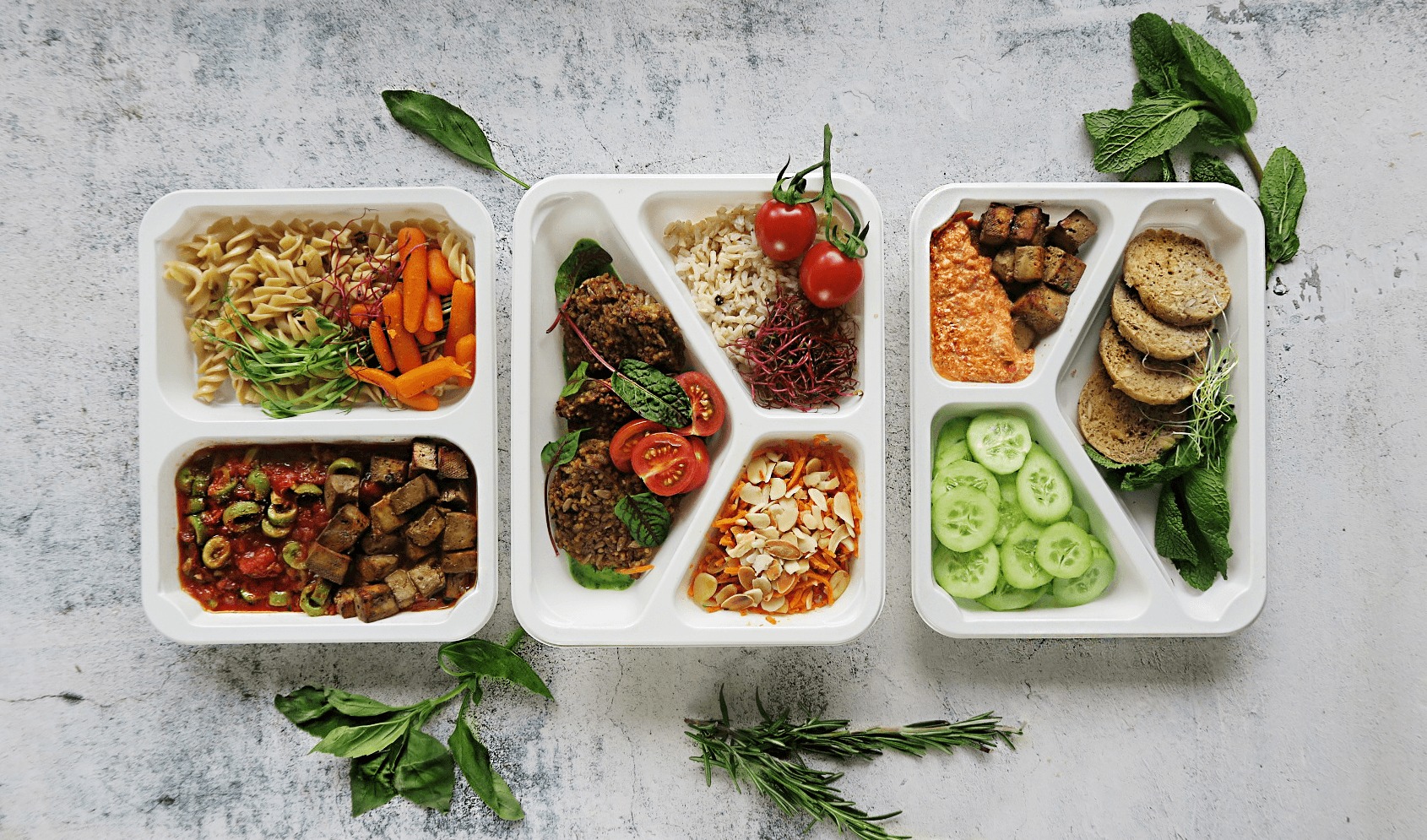 Trzy białe tacki z podziałem na komory, zawierające gotowe dania: makaron z warzywami, kotleciki z kaszy z warzywami, pieczywo z warzywami i dipem, całość uzupełniona świeżymi ziołami, na szarym tle.