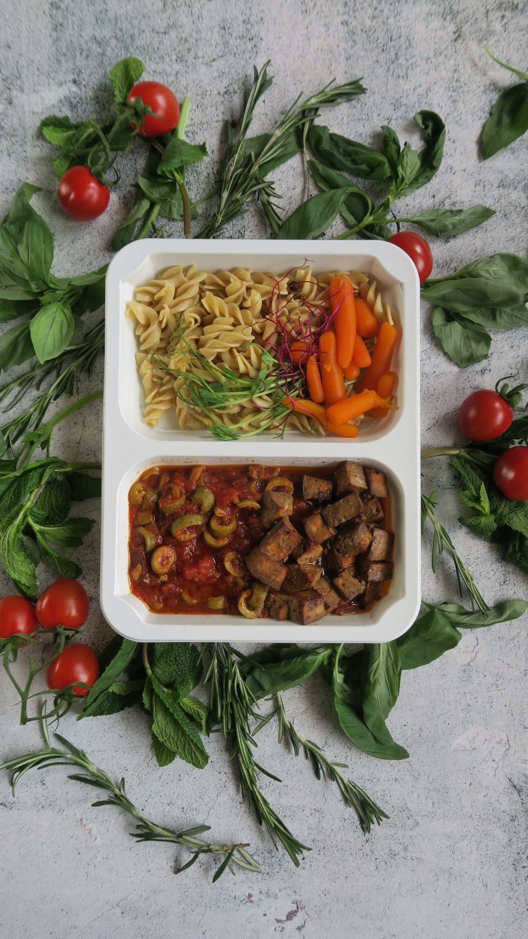 Dwu-komorowe pudełko z gotowym posiłkiem: makaron świderki z zielonymi pędami i marchewką baby oraz gulasz z tofu z oliwkami w sosie pomidorowym, otoczone świeżymi ziołami i pomidorkami cherry...