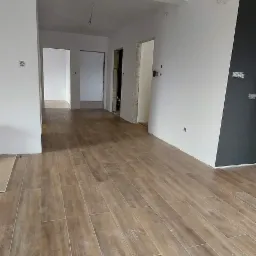 Ułożona podłoga z płytek imitujących drewno w przestronnym, częściowo wykończonym wnętrzu. Widoczne surowe ściany, niezamaskowane kable i otwarte przejścia do innych pomieszczeń.