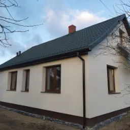 Dom jednorodzinny po remoncie elewacji, z nowym, ciemnozielonym dachem, brązowymi oknami i orynnowaniem, na tle pochmurnego nieba i drzew bez liści.