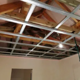 Montaż metalowego stelaża sufitu podwieszanego z płyt karton gips, widoczny laserowy poziom na ścianie, oświetlenie robocze i elementy konstrukcji drewnianej dachu.