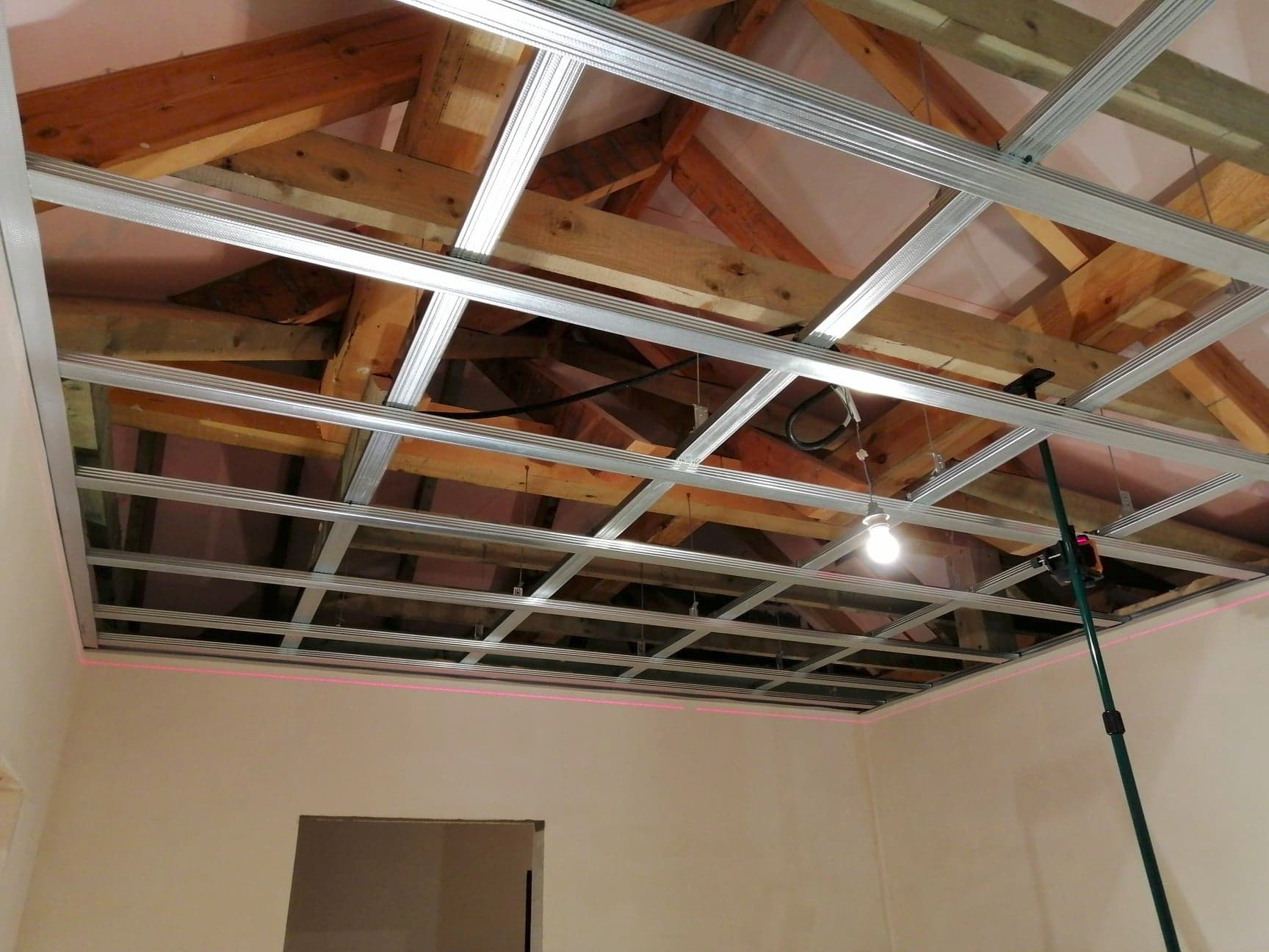 Montaż metalowego stelaża sufitu podwieszanego z płyt karton gips, widoczny laserowy poziom na ścianie, oświetlenie robocze i elementy konstrukcji drewnianej dachu.