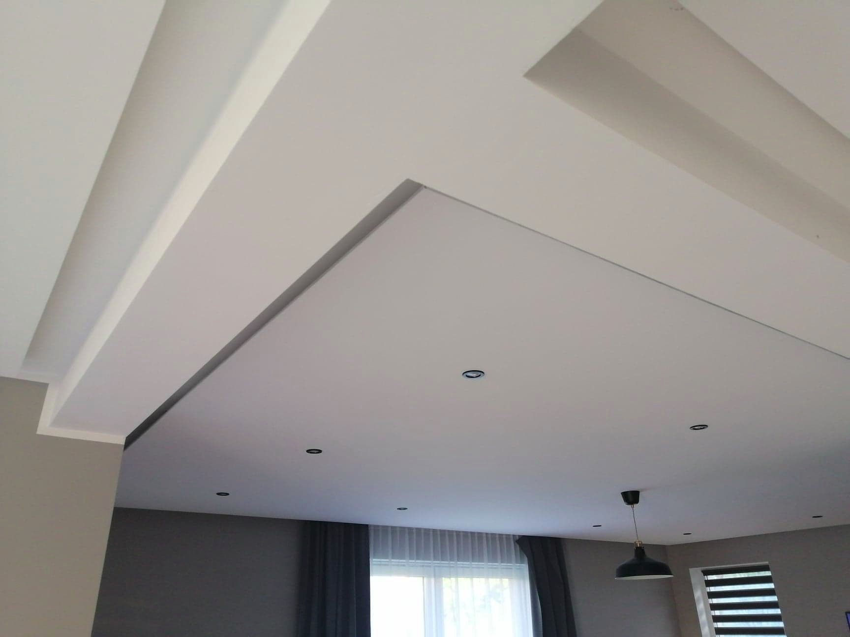 Nowoczesny sufit podwieszany z oświetleniem punktowym i dekoracyjnymi listwami, widok z dołu, szare ściany i zasłony w tle.