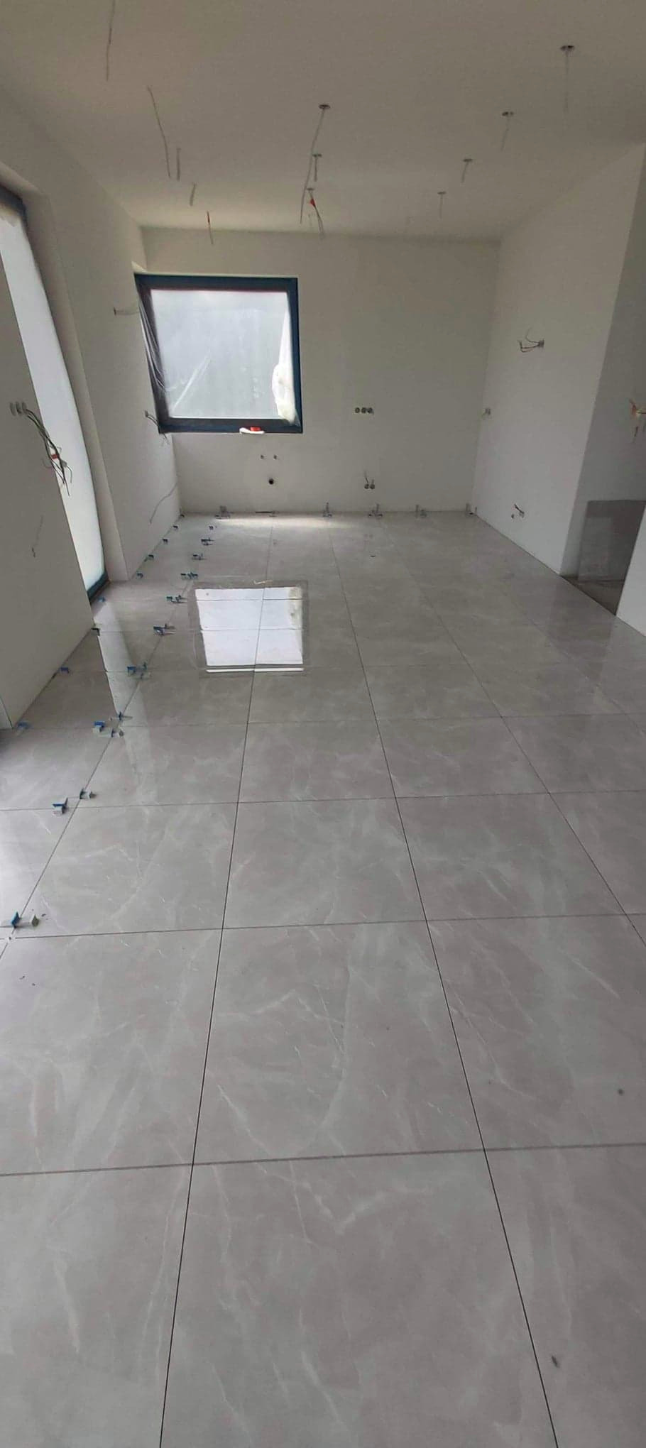 Jasnoszare płytki podłogowe imitujące marmur, ułożone w przestronnym, jasnym pomieszczeniu z oknem dachowym, widoczne plastikowe krzyżyki dystansowe